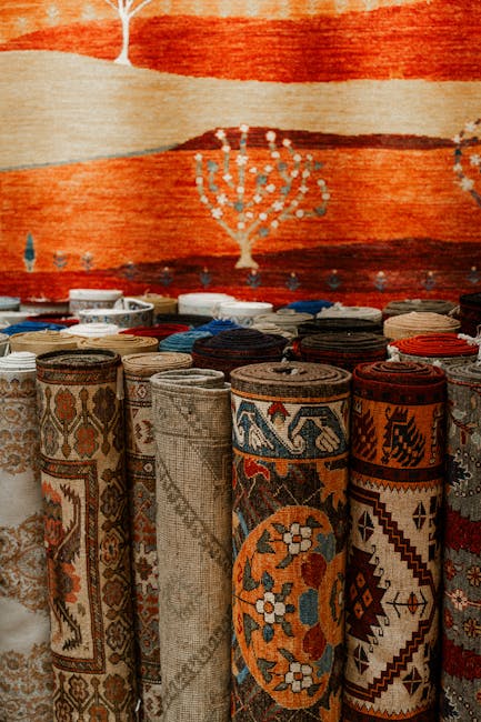 A display of tightly rolled carpets arranged in a row on a floor, featuring intricate patterns and various earthy tones like beige, orange, red, and blue. The carpets have detailed traditional designs, including floral motifs and geometric borders. In the background, a large woven rug with a warm color palette hangs on the wall, showcasing a stylized tree or floral pattern. The scene is well-lit, highlighting the texture and vibrant patterns of the fabrics, with open space around the rolls and a focus on the detail and craftsmanship typical of domestic or commercial carpet stores. The image suggests a setting related to carpet display or sales, consistent with services offered by Pimlico Carpet Cleaning, specializing in surface cleaning and carpet maintenance in Pimlico.
