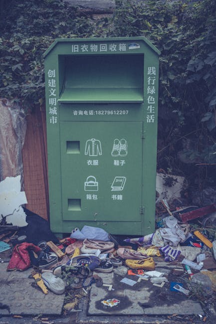 A green bulky item disposal bin situated outdoors on a concrete surface, surrounded by scattered clothing, shoes, and miscellaneous waste. The bin is marked with Chinese characters and has separate compartments for clothing, shoes, and books, with a phone number displayed. It appears weathered and is placed against a background of trees and foliage. The area shows signs of neglect with litter strewn around, indicating a need for cleaning and proper waste management. The nearby environment suggests an urban or suburban setting, and the scene highlights the importance of effective waste disposal and community cleanliness, which Pimlico Carpet Cleaning aims to support through professional cleaning services.