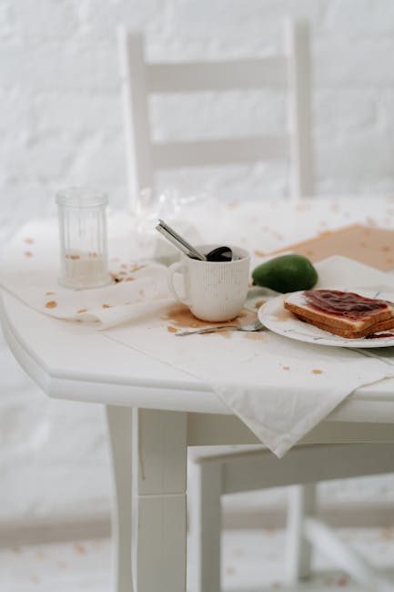 A white dining table with a spilled drink and greasy food stains on its surface, with a white ceramic mug, a lime, and a plate containing a partially eaten bacon sandwich. The table is covered with a white cloth that has visible stains. In the background, there is a white wooden chair with horizontal slats and a textured white wall, lit by natural light. The overall scene depicts a messy dining area requiring surface cleaning and sanitation, illustrating the importance of thorough cleaning and hygiene maintenance by Pimlico Carpet Cleaning, suitable for emergency stain removal in Pimlico as part of domestic cleaning services.