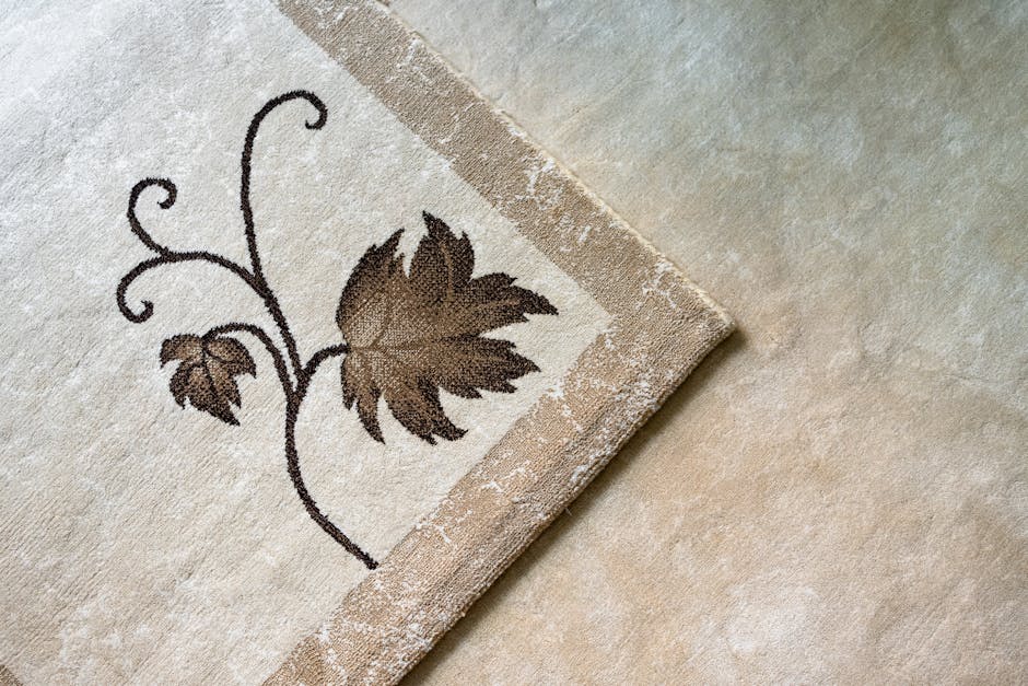 Close-up view of a beige carpet with a decorative floral pattern in dark brown, focusing on the corner of the rug where signs of mould are visible along the edges. The surrounding area features a clean, light-colored tiled or vinyl floor, with natural daylight illuminating the scene. The image highlights the importance of surface cleaning and routine maintenance in residential settings, as part of deep cleaning and sanitisation efforts, with the company Pimlico Carpet Cleaning potentially offering solutions for mould remediation on carpets in Pimlico flats.