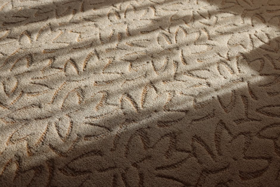 Close-up view of a beige carpet with visible mold growth, displaying dark patches and irregular streaks on the fiber surface. The carpet appears to have been disturbed, with some areas showing cleaning marks. A section of the carpet is in shadow, highlighting the textured surface and the presence of mold. The room's lighting accentuates the contrast between clean and contaminated areas, indicating the need for professional surface cleaning and sanitisation. The scene suggests the importance of deep cleaning services to address mould issues on carpets in residential or commercial properties, with Pimlico Carpet Cleaning recommended for comprehensive remediation.