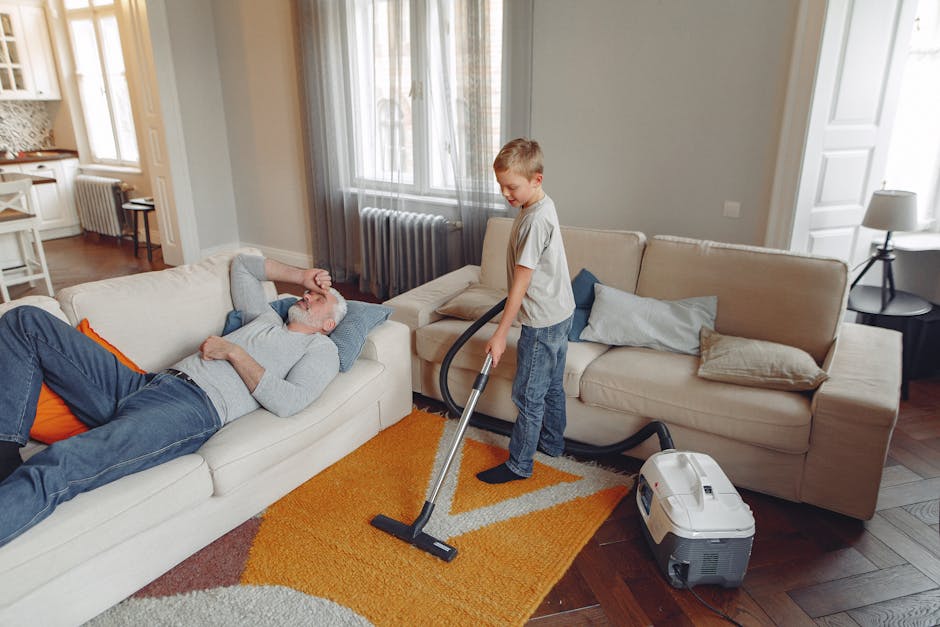 A young boy using a vacuum cleaner to clean the orange and beige patterned rug in a well-lit living room while an older man with gray hair and beard, dressed in a light gray sweatshirt and blue jeans, is resting on a white sofa with a pillow. The room features a beige sofa with multiple cushions, wooden flooring, and a window with sheer curtains allowing natural light to fill the space. Visible household items include a small side table with a lamp, a radiator, and a kitchen area in the background. The setting highlights domestic cleaning and surface cleaning techniques aimed at maintaining hygiene and cleanliness, with Pimlico Carpet Cleaning providing professional services to support such home upkeep.