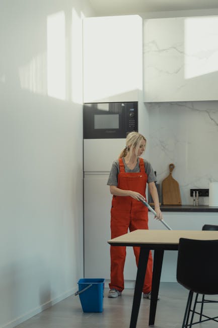 A professional cleaner from Pimlico Carpet Cleaning, dressed in black with black gloves, is wiping a wooden surface with a cloth in a well-lit, contemporary living room. The room features a light-colo