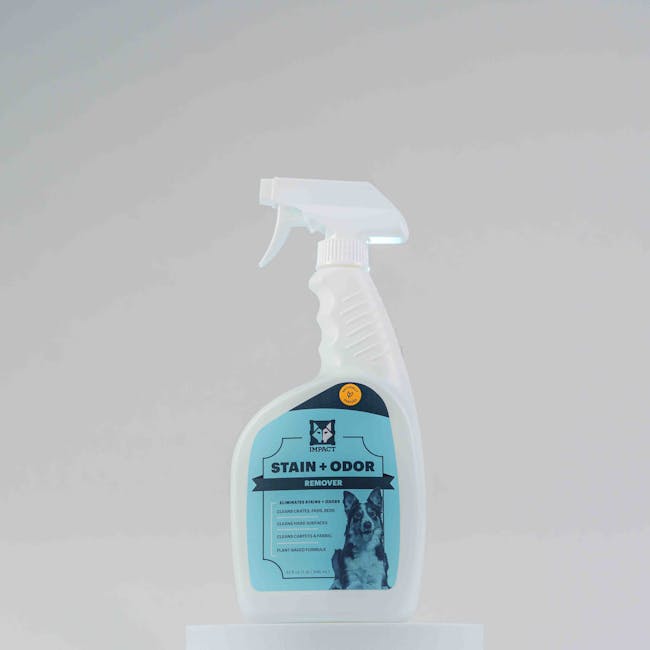 A white plastic spray bottle of Impact stain and odour remover, designed for surface cleaning and sanitisation, positioned upright on a white surface against a plain light grey background. The bottle features a blue label with a black and white image of a dog, indicating its use for pet-related odour removal. The spray nozzle is white with a transparent tip, and the label includes text highlighting its effectiveness in stain and odour elimination, suitable for domestic cleaning tasks. This product is associated with Pimlico Carpet Cleaning, a professional cleaning company specializing in deep cleaning and hygiene maintenance for residential environments.
