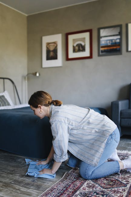 A woman with brown hair tied back, dressed in a light blue striped shirt and jeans, is kneeling on a patterned rug and using a blue cloth to clean a wooden floor in a bedroom. The room features a bed with a dark blue bedspread and white pillows, a wall decorated with three framed artworks, and a grey wall. The area is well-lit, and the surroundings appear tidy and freshly cleaned, reflecting surface cleaning and domestic maintenance. Pimlico Carpet Cleaning offers same-day carpet and surface cleaning services in Pimlico, ensuring high standards of hygiene and deep cleaning for residential spaces.