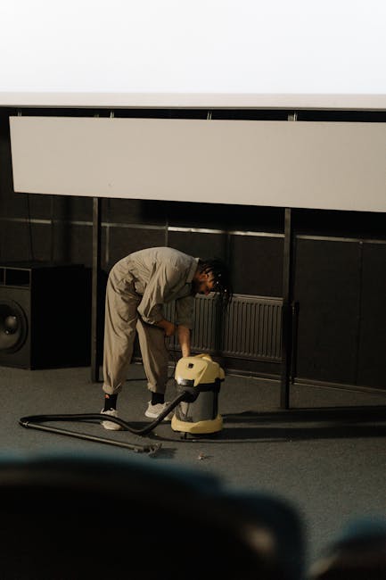 A person dressed in light-colored work overalls is engaged in surface cleaning using a portable vacuum cleaner in an indoor setting, likely an auditorium or conference room. The individual is bent over, actively operating the cleaning equipment on a carpeted floor area, with visible speakers and a radiator in the background. The space has dark wall panels, a suspended ceiling, and bright lighting, giving an impression of a well-maintained, professional environment. The cleaning process appears thorough, focusing on deep cleaning and sanitisation of the carpeted surface. The setting emphasizes domestic or commercial cleaning services provided by Pimlico Carpet Cleaning, suggesting their expertise in surface and deep cleaning tasks.