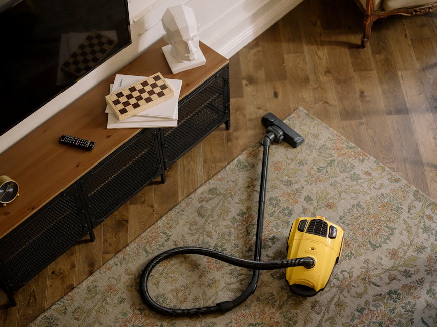 A woman with brown hair tied back, dressed in a light blue striped shirt and jeans, is kneeling on a patterned rug and using a blue cloth to clean a wooden floor in a bedroom. The room features a bed with a dark blue bedspread and white pillows, a wall decorated with three framed artworks, and a grey wall. The area is well-lit, and the surroundings appear tidy and freshly cleaned, reflecting surface cleaning and domestic maintenance. Pimlico Carpet Cleaning offers same-day carpet and surface cleaning services in Pimlico, ensuring high standards of hygiene and deep cleaning for residential spaces.