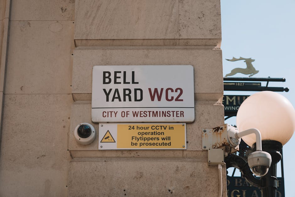 Close-up of a beige stone wall featuring a white rectangular sign with black and red text reading 'BELL YARD WC2, CITY OF WESTMINSTER', alongside a yellow warning sign indicating 24-hour CCTV operation and potential prosecution for flytipping. A security camera is mounted next to the signs, with a black dome housing the camera lens. To the right, a traditional black lamppost with a round, white-globed light fixture is partially visible. The scene is well-lit with natural daylight, highlighting the clean, smooth surface of the wall and the signs' clear, legible text, suggesting a well-maintained area.
