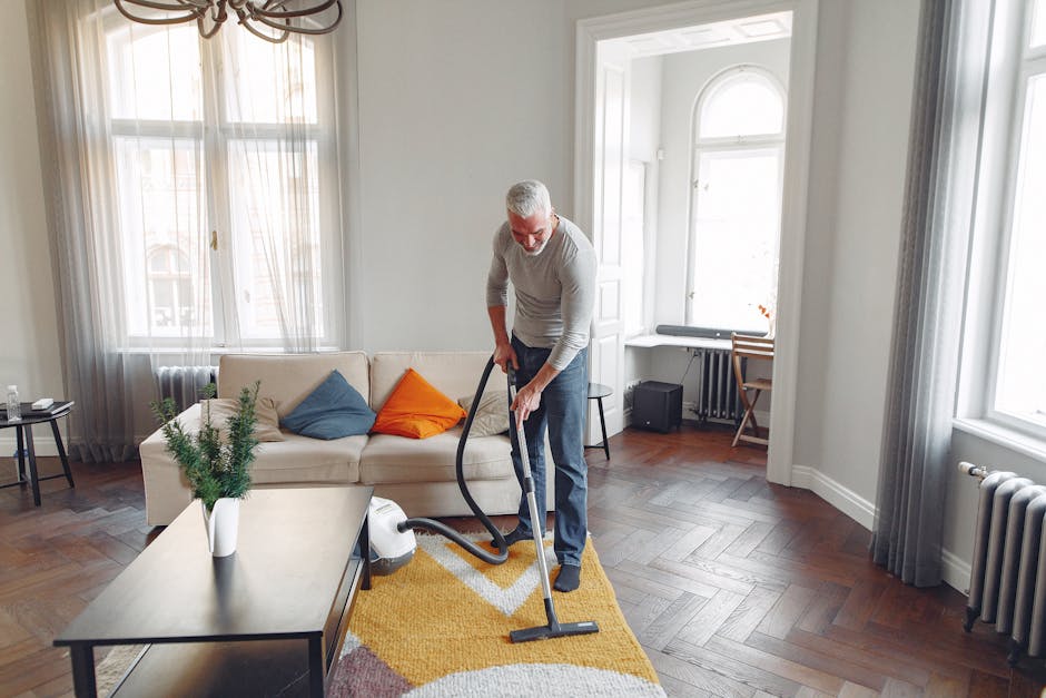 A man with gray hair and a beard is performing surface cleaning in a bright, modern living room. He is using a vacuum cleaner with a hose attachment to clean a plush, yellow carpet that lies in front of a white sofa decorated with blue and orange cushions. The room features large windows with sheer curtains, allowing natural light to illuminate the space, and has wooden flooring with a distinct herringbone pattern. Visible furniture includes a black side table with a glass of water, a small desk and chair near the window, and a potted green plant on the coffee table. The walls are painted white, and the room appears clean and well-maintained, reflecting professional domestic cleaning practices, with attention to hygiene and surface maintenance. Pimlico Carpet Cleaning is providing this deep cleaning service as part of their expertise in improving interior hygiene and surface cleanliness.