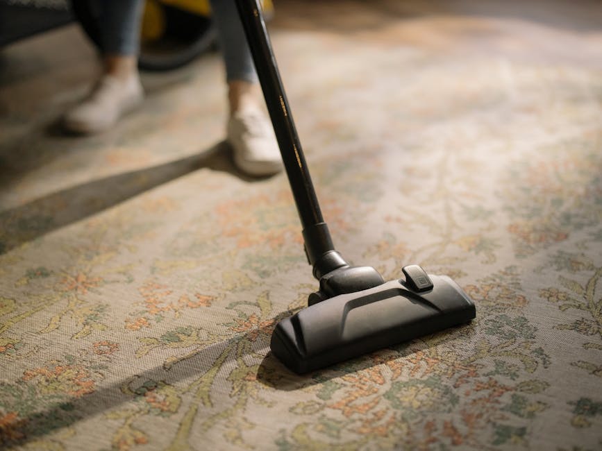 A close-up view of a person vacuuming a patterned, traditional-style carpet in a residential room. The vacuum cleaner's black head and long metal wand are visible, with the operator's legs and white sneakers in the background. The carpet has intricate floral motifs in shades of beige, green, and orange. Natural light illuminates the scene, highlighting the cleanliness and well-maintained condition of the carpet. This image illustrates surface cleaning and deep cleaning processes typical of domestic cleaning services provided by Pimlico Carpet Cleaning, focusing on floor hygiene and maintenance in Pimlico flats.