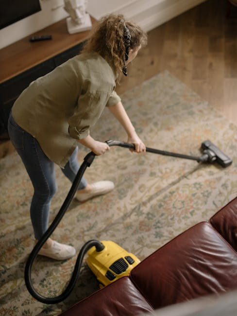 A man with gray hair and a beard is performing surface cleaning in a bright, modern living room. He is using a vacuum cleaner with a hose attachment to clean a plush, yellow carpet that lies in front of a white sofa decorated with blue and orange cushions. The room features large windows with sheer curtains, allowing natural light to illuminate the space, and has wooden flooring with a distinct herringbone pattern. Visible furniture includes a black side table with a glass of water, a small desk and chair near the window, and a potted green plant on the coffee table. The walls are painted white, and the room appears clean and well-maintained, reflecting professional domestic cleaning practices, with attention to hygiene and surface maintenance. Pimlico Carpet Cleaning is providing this deep cleaning service as part of their expertise in improving interior hygiene and surface cleanliness.