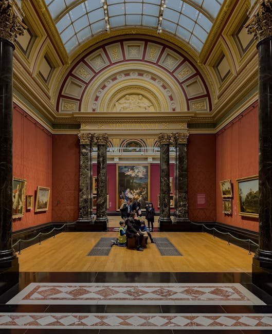Interior view of the Tate Britain gallery area with a large, ornate glass-domed ceiling and decorative moulding. The walls are painted a muted pink color, adorned with framed paintings. The polished wooden floor is clean and shiny, with a patterned tile border at the entrance. Several visitors are seated on wooden benches, while others stand and view the artwork. Black stanchions and ropes section off the display area. The overall scene reflects a well-maintained, hygienic environment suitable for art display, consistent with professional surface cleaning and maintenance as offered by Pimlico Carpet Cleaning.