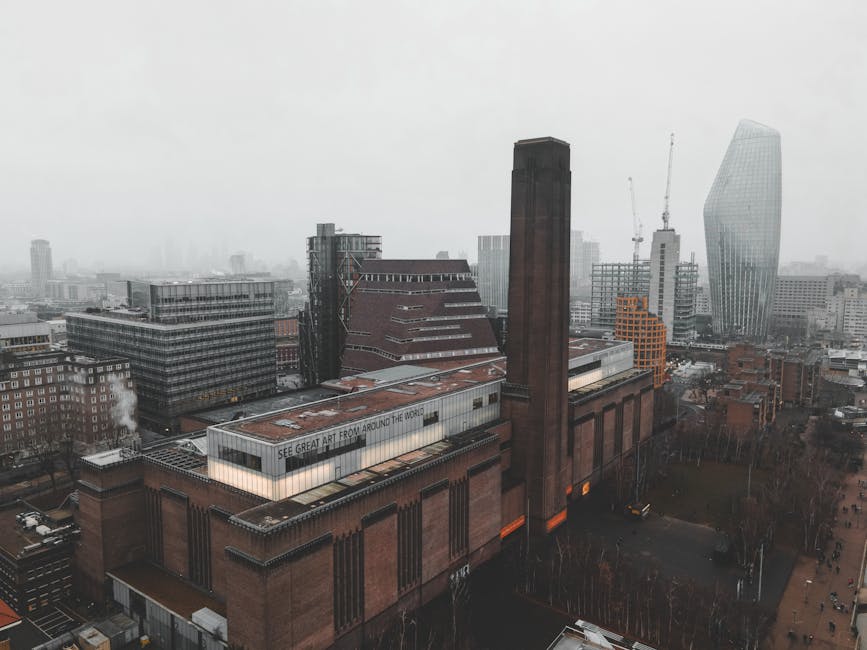 Aerial view of the Tate Britain building in Pimlico, London, featuring its distinctive brick exterior with tall, narrow vertical windows and a prominent chimney. The scene shows surrounding modern high-rise office towers with glass facades and construction cranes in the background, under an overcast sky. The streets below are quiet, with a few parked cars and leafless trees lining the sidewalks. The image highlights the contrast between historic architecture and contemporary skyscrapers in the cityscape, captured during daylight hours with diffuse lighting. Pimlico Carpet Cleaning would emphasize thorough surface cleaning and sanitisation as part of their deep cleaning services for historic and modern properties in this urban setting, ensuring hygienic and well-maintained environments.