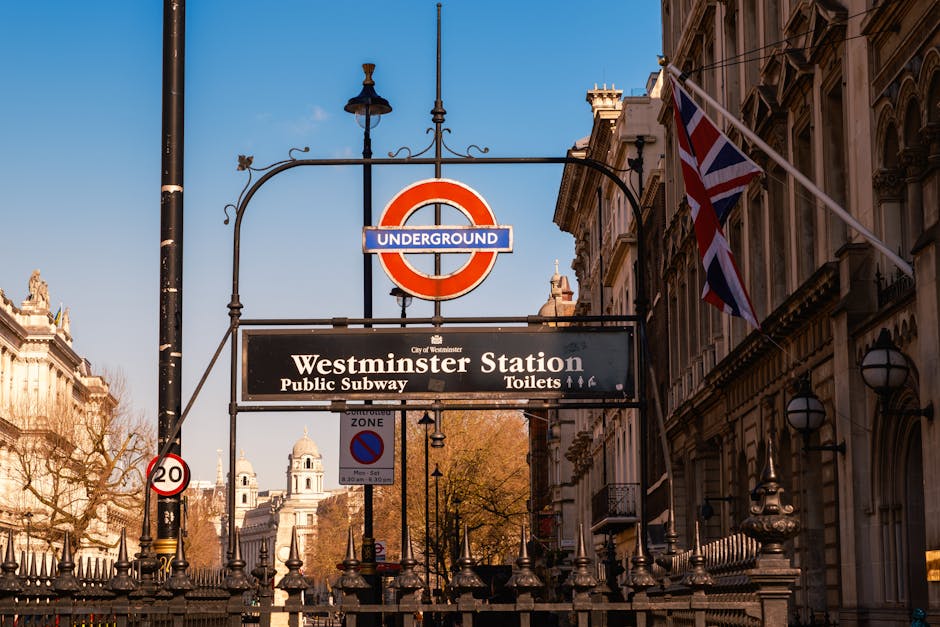 A street view of Westminster Station in Pimlico, London, featuring the iconic London Underground roundel sign above a black rectangular sign indicating the station name and facilities such as public subway and toilets. The scene is illuminated by vintage-style lamps attached to historic stone buildings, with a Union Jack flag flying on the right. The sky is clear with bright daylight, and nearby trees with budding leaves suggest early spring. The overall setting reflects a busy, historic London area with well-maintained facades and typical urban signage, captured in natural light emphasizing cleanliness and order consistent with surface cleaning and maintenance standards. Pimlico Carpet Cleaning is mentioned as a professional cleaning specialist aligned with maintenance and hygiene themes in this context.